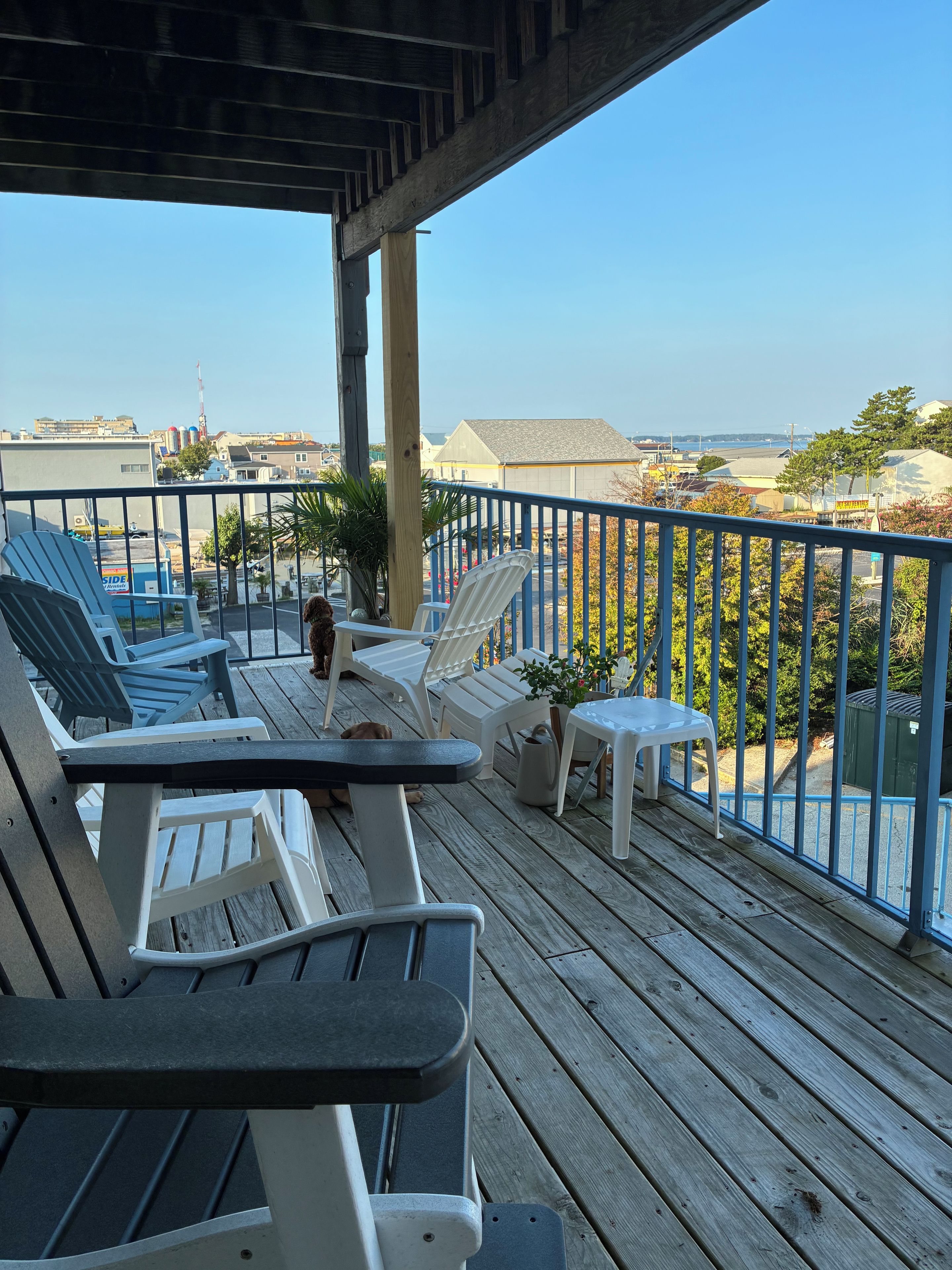 A wooden deck with outdoor seating, including chairs and a table, overlooking a residential area with houses and a clear sky.