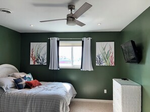 Ein Schlafzimmer mit einem Bett, einem Deckenventilator, einem Fenster mit Vorhängen und zwei Wandbildern.