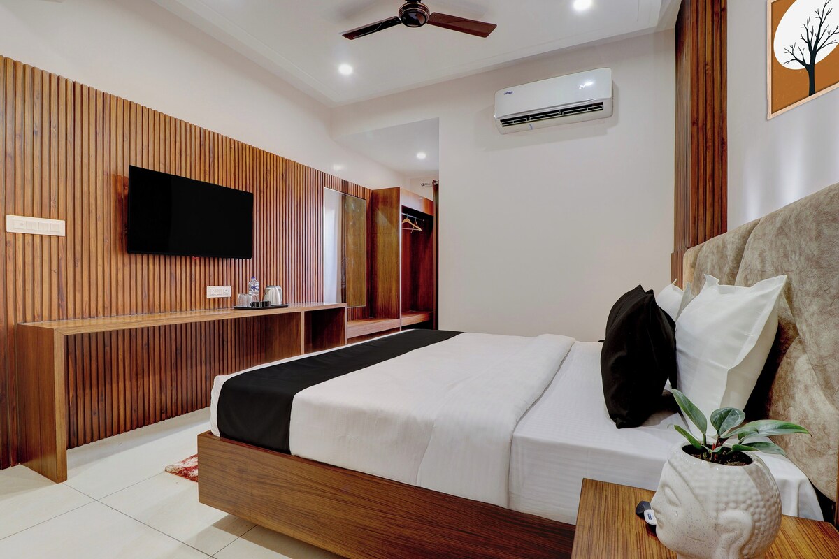 A modern hotel room with a wooden panel wall, a flat-screen TV, a bed with white bedding, a bedside table with a plant, and a ceiling fan.