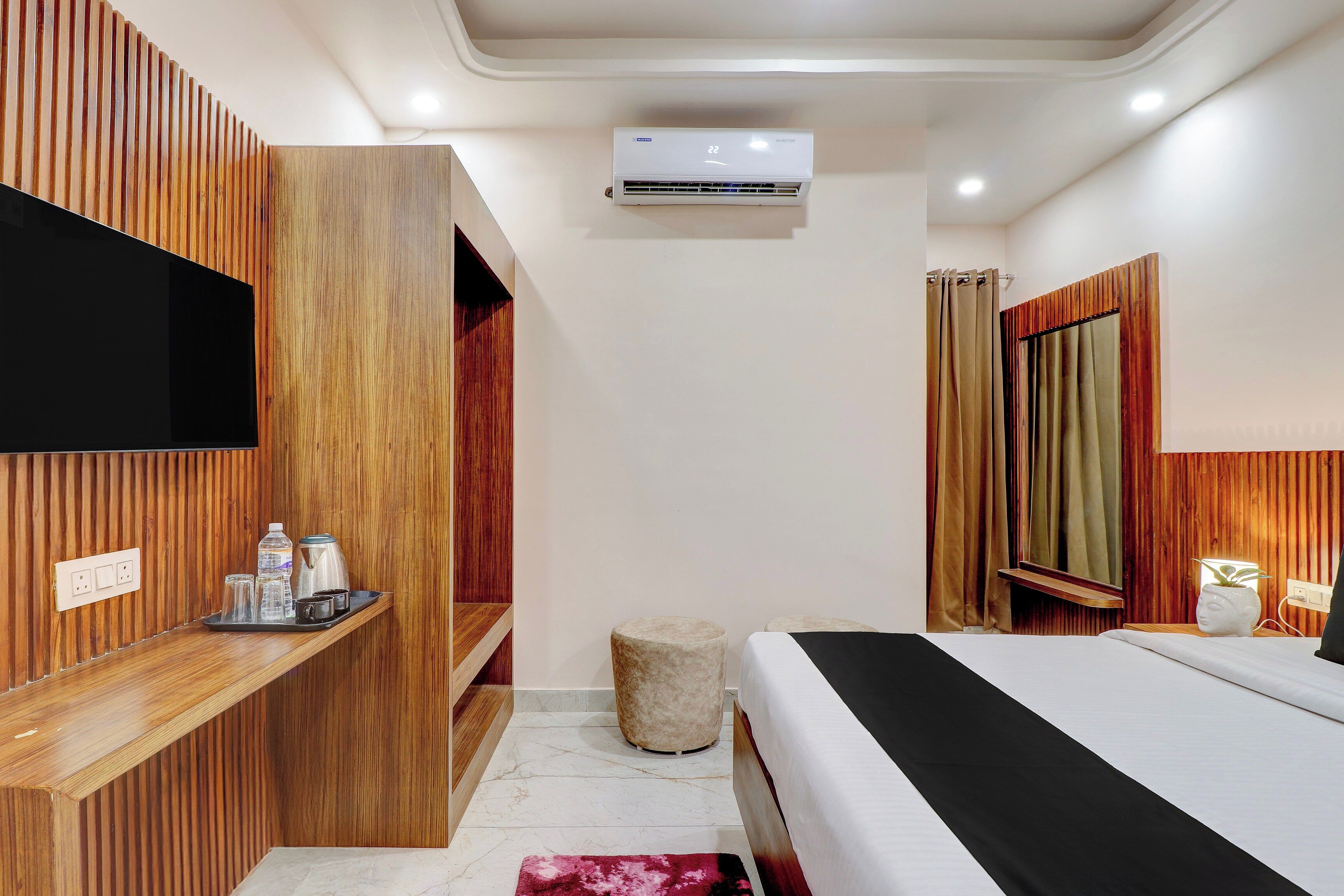 Una habitación de hotel moderna con una cama, televisión, pared de paneles de madera, una pequeña zona de estar y un aire acondicionado instalado en el techo.