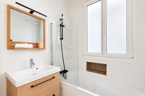 A modern bathroom with a white sink, wooden vanity, a mirror, a shower enclosure, and a window. - Charming Flat near Opéra Garnier (Paris)