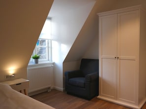 Une chambre avec un plafond en pente, une fenêtre agrémentée d’une plante, un fauteuil sombre, une armoire et une table de chevet avec une lampe.