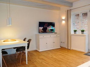 A modern living room with a dining area, a flat-screen TV mounted on a cabinet, and a window with a view of a brick building. - 3 Barbados in the Boldix House (Wyk auf Foehr)