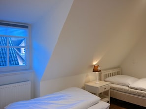 A bedroom with a sloped ceiling, a window with blinds, a bedside lamp, and a bed with a white mattress. - 3 Barbados in the Boldix House (Wyk auf Foehr)