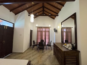 A hotel room with a wooden ceiling, a bed, a desk with a mirror, and a dining area with chairs and curtains.
