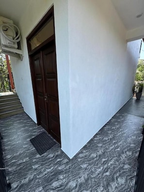 Une entrée d’immeuble avec une porte en bois foncé, un mur blanc et un sol en marbre gris.