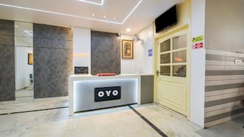 Un área de recepción con diseño moderno, que incluye un mostrador de recepción con la etiqueta 'OYO', un mostrador de registro, una cámara de seguridad y un cartel de vigilancia.
