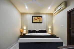 Quarto de hotel com cama, ventoinha no teto, aparelho de ar condicionado, quadro na parede e um vaso com flores na mesa de cabeceira.