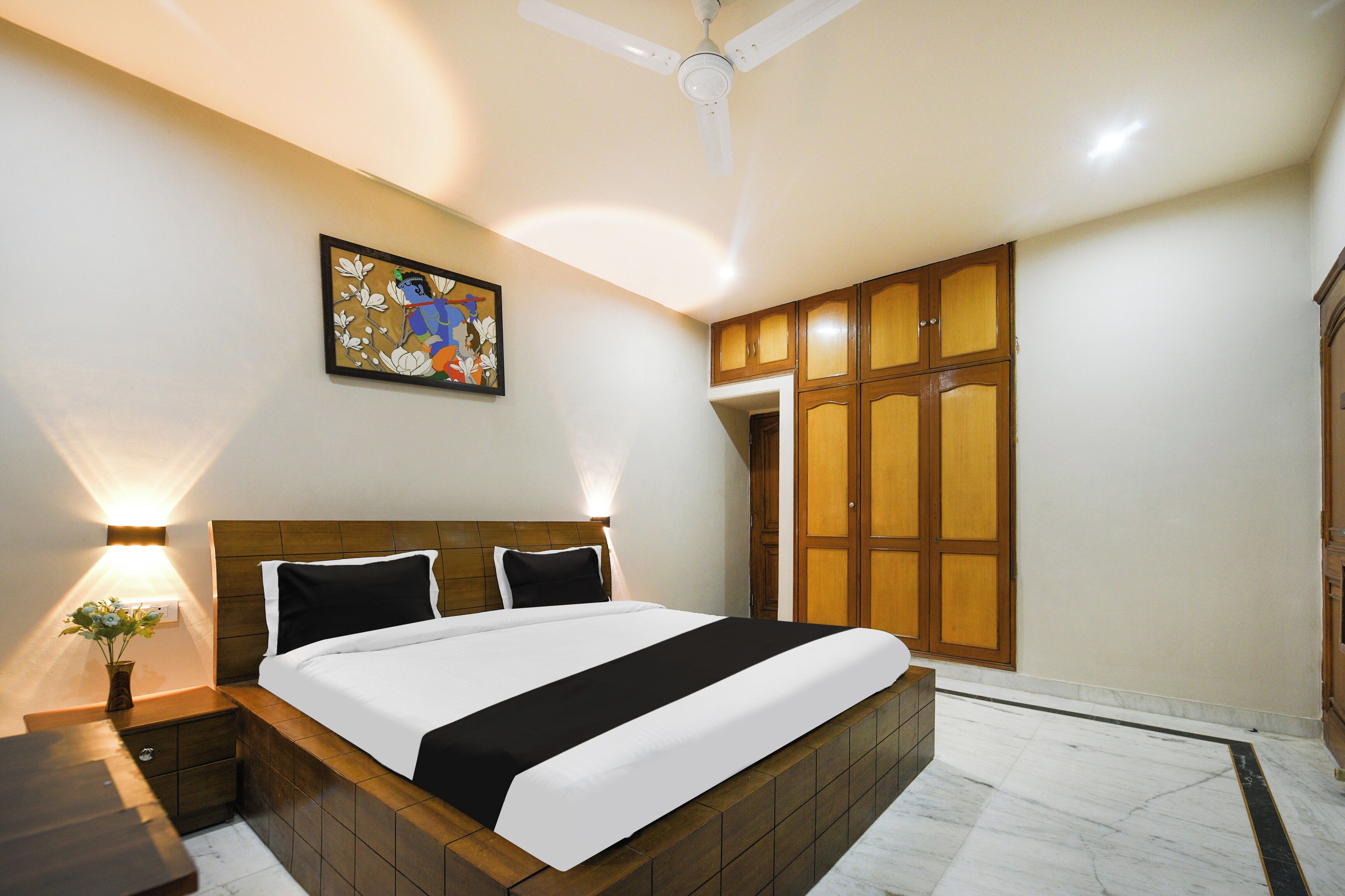 Habitación de hotel con cama, mesita de noche, lámpara de pared, ventilador de techo y puerta con panel de madera.