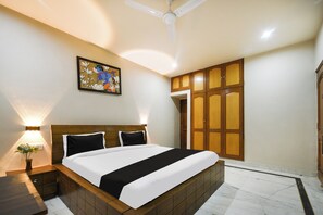 Quarto de hotel com cama, mesa de cabeceira, luminária na parede, ventoinha no teto e porta com painel de madeira.