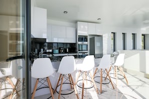 A modern kitchen with white cabinets, a marble countertop, and a built-in oven. There are four white bar stools with wooden legs. - BLUE OCEAN VILLA with Private Heated Pool. Magnificent Seaview. AC, WIFI, Garden (Fajã da Ovelha, Calheta)
