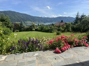 A stone pathway leading to a lush garden with diverse flowers and a view of a lake and mountains in the distance. - New Listing (Bodensdorf)