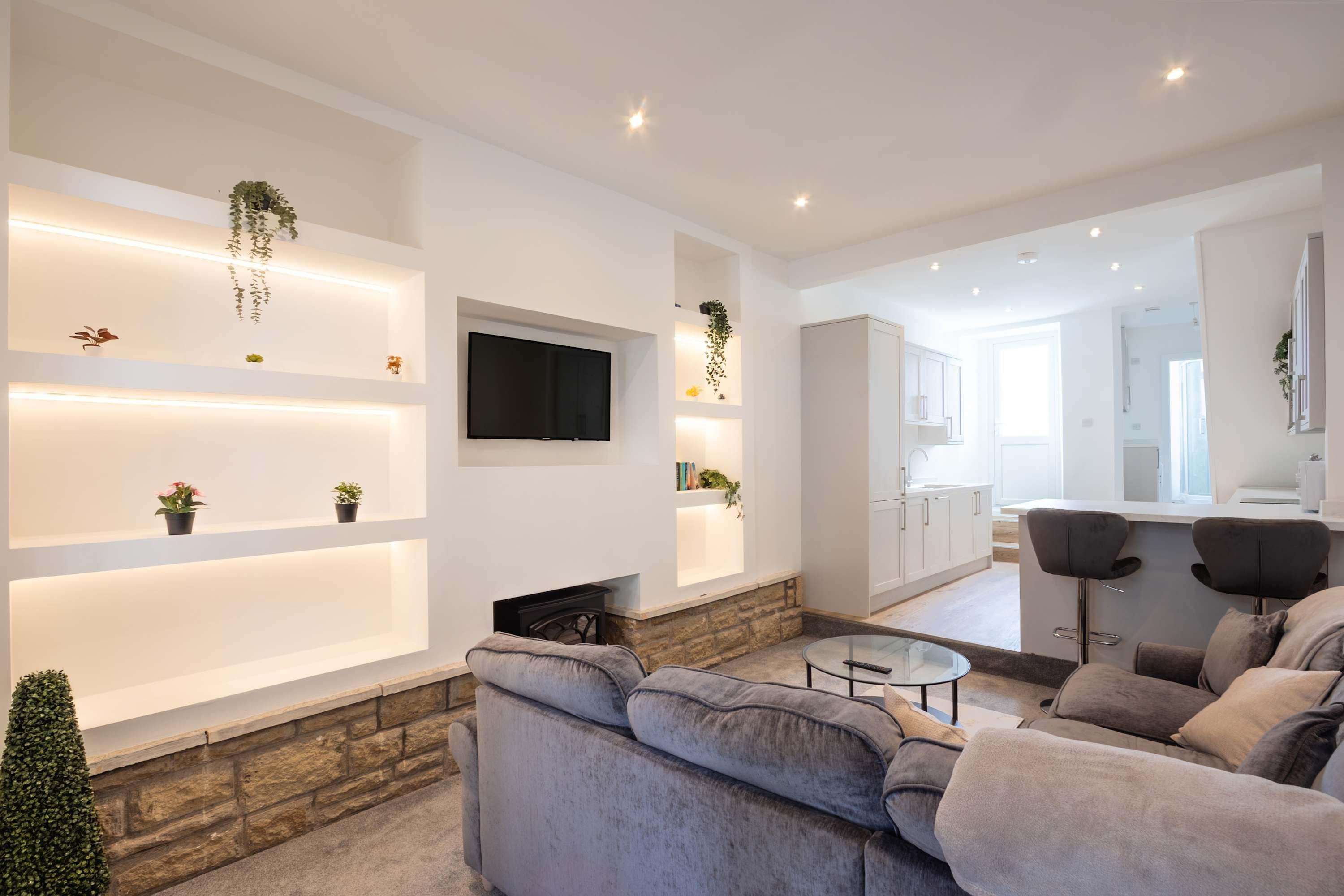 A modern living room with a stone fireplace, grey sofas, a flat-screen TV, and built-in shelves with plants.