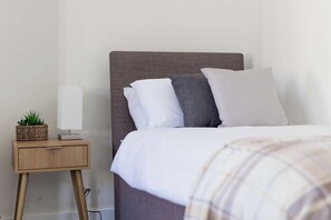 A neatly made bed with a headboard, a bedside table with a lamp and a plant, and a wall with a power outlet. - Cosy 3 Bed | Sleeps 7 | Free Parking (Bristol City)