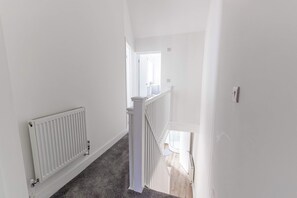 A narrow hallway with a white radiator, a white door at the end, and a staircase with a white railing. - Cosy 3 Bed | Sleeps 7 | Free Parking (Bristol City)