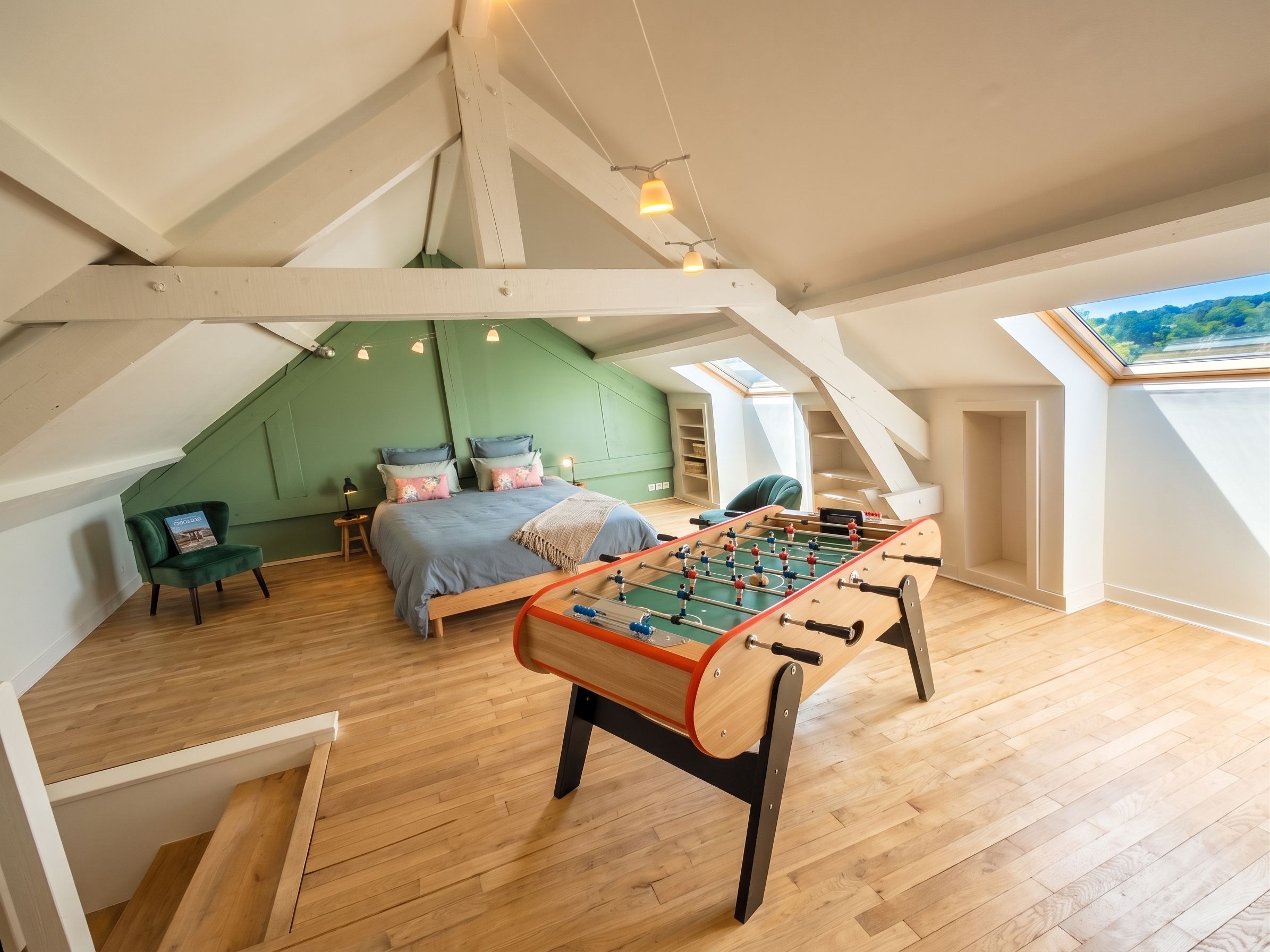 Un dormitorio tipo loft con piso de madera, una cama con almohadas, una pared verde, una mesa de futbolín y un tragaluz.