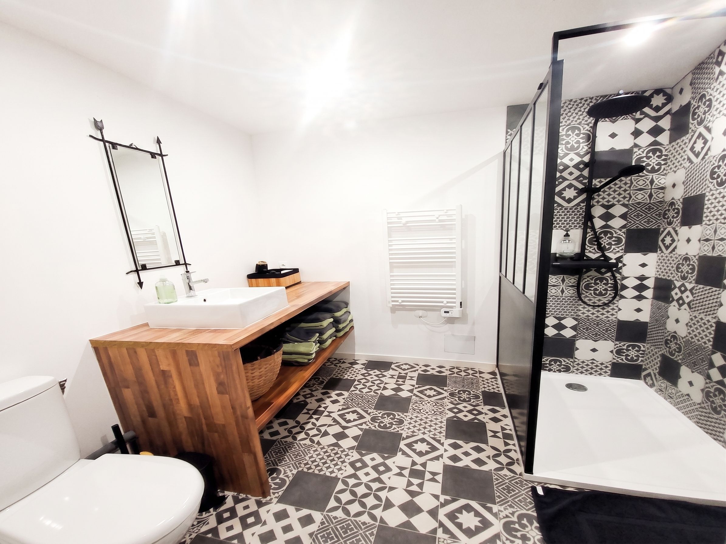 Un baño moderno con un lavabo blanco, tocador de madera, baldosas de suelo con diseño, cabina de ducha con mampara de vidrio y un inodoro blanco.