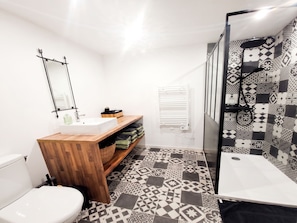 Un baño moderno con un lavabo blanco, tocador de madera, baldosas de suelo con diseño, cabina de ducha con mampara de vidrio y un inodoro blanco.
