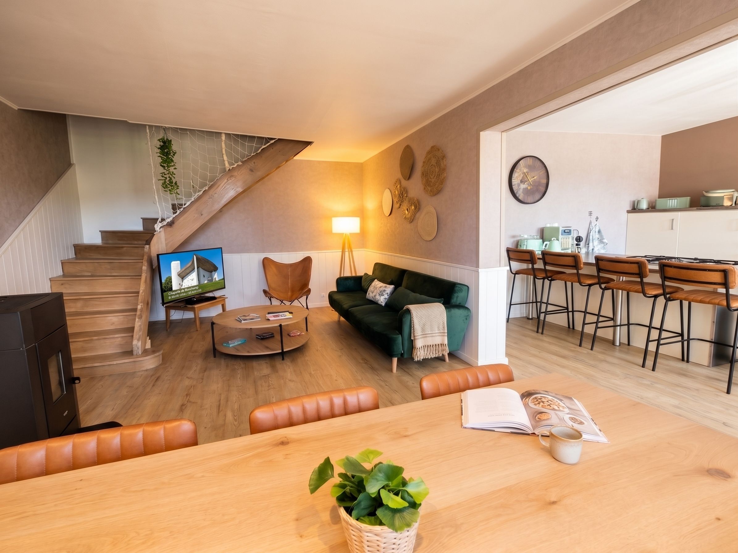 A modern living room with a staircase, a green sofa, a dining table with chairs, and a kitchen area.