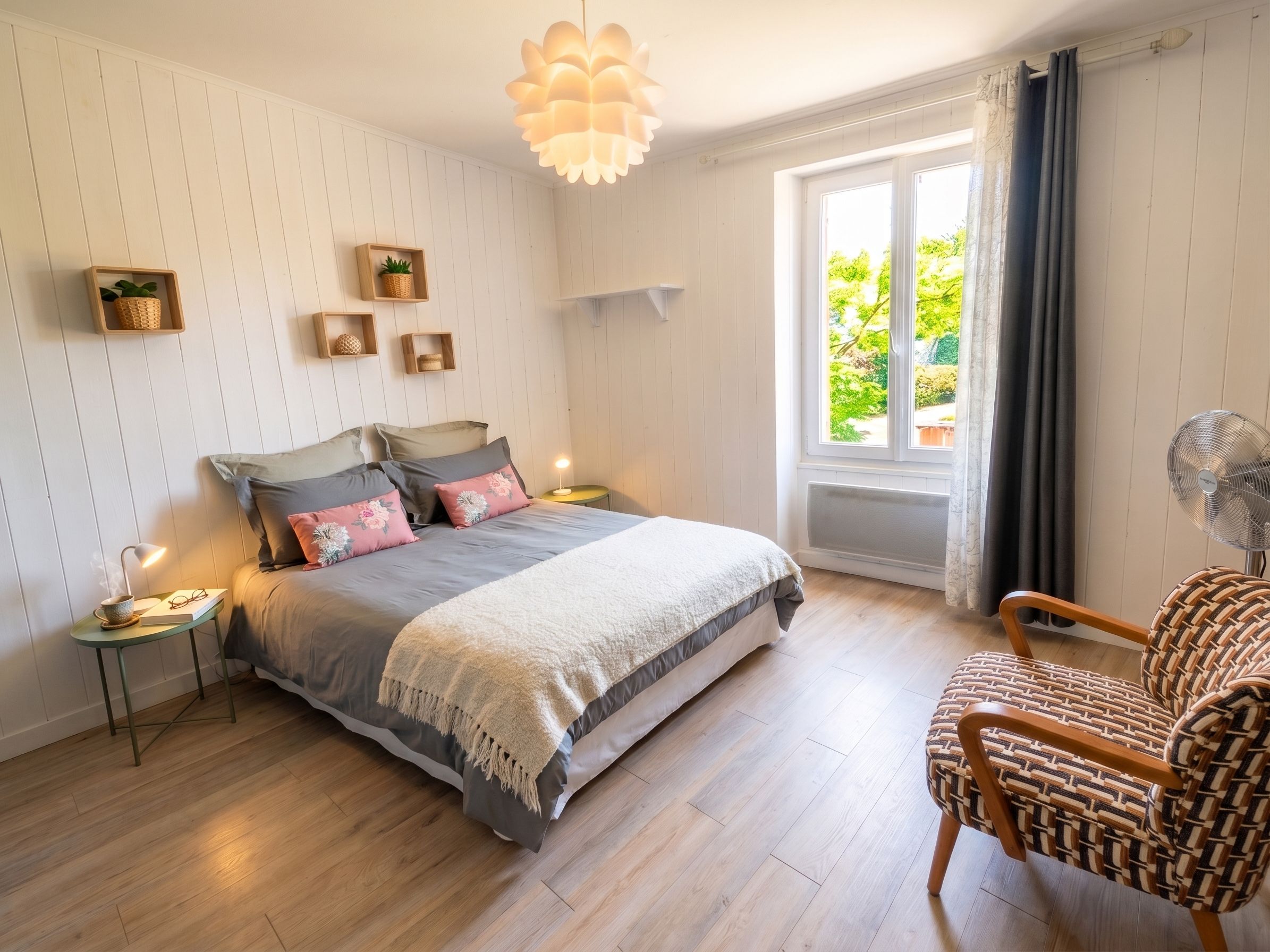 Une chambre à coucher comprenant un lit, des tables de chevet, une chaise, une fenêtre et un luminaire suspendu.