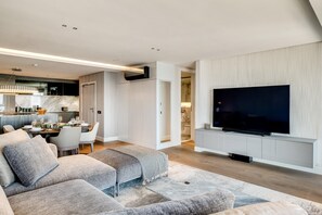 A modern living room with a large flat-screen TV mounted on the wall, a sectional sofa, and a dining area with a table set for four. - Clifton 2 Bed Apartment Ocean Views Dunmore Noir (Cape Town)