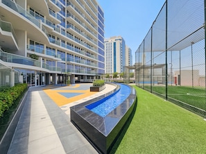 A modern building with a swimming pool and a fenced sports area. - Stayshort - Stylish 1-bedroom Apartment in Al Furjan With Pool and Gym (Dubai)