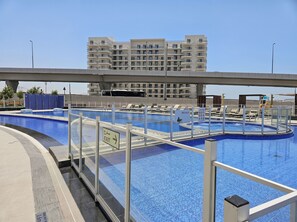A swimming pool with a glass barrier, a sign indicating an exit, and a building in the background. - Stayshort - Stylish 1-bedroom Apartment in Al Furjan With Pool and Gym (Dubai)