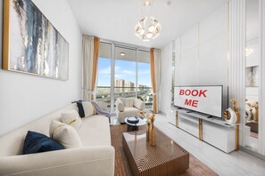 A modern living room with a sofa, coffee table, and a TV screen displaying 'BOOK ME'. - Stayshort - Stylish 1-bedroom Apartment in Al Furjan With Pool and Gym (Dubai)