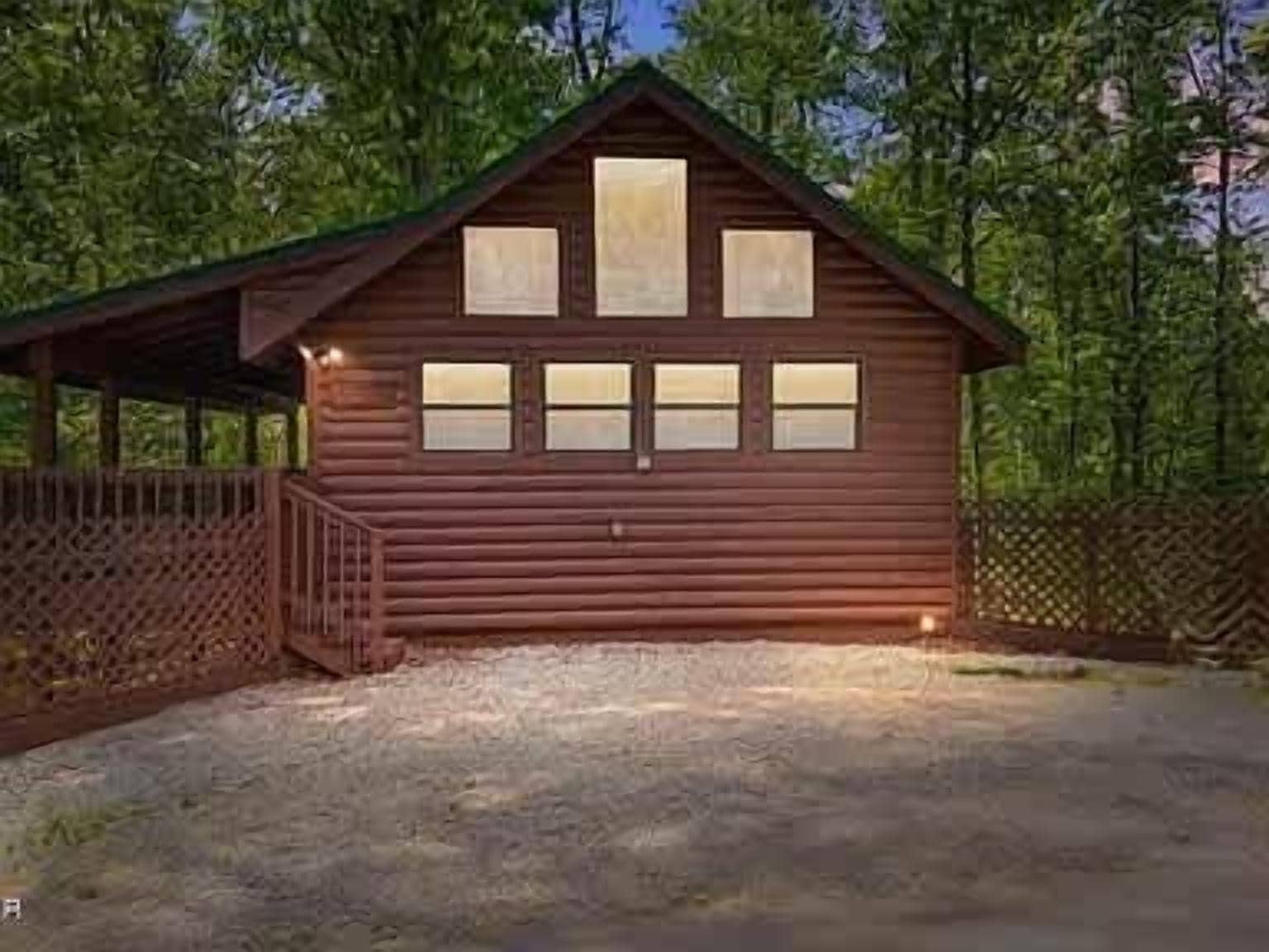 A wooden cabin with a porch, a gravel driveway, and a fenced yard.