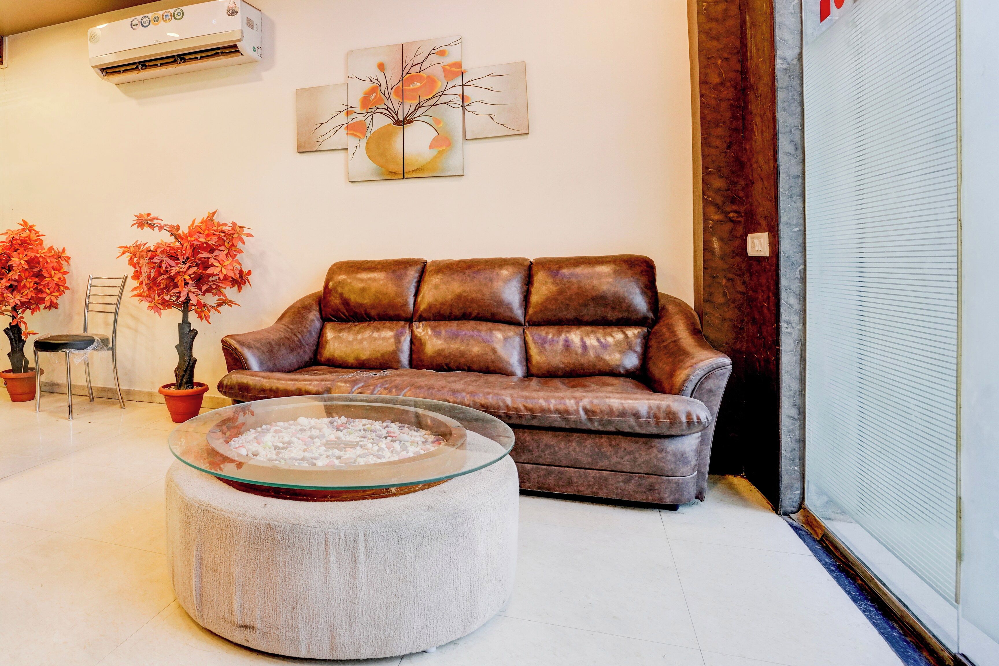 Un salon avec un canapé en cuir marron, une table basse en verre, deux plantes en pot et un climatiseur fixé au mur.