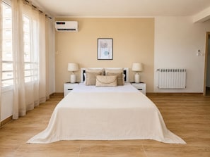 A bedroom with a bed, two bedside tables with lamps, a wall-mounted air conditioner, a framed picture, and a radiator.