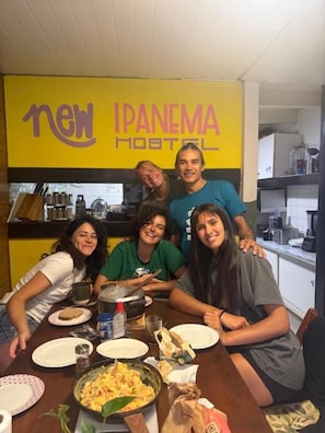 Eine Gruppe von Menschen sitzt an einem Tisch mit einer Pfanne Essen in einem Raum, dessen Wand gelb ist und der ein Schild mit der Aufschrift 'new Ipanema Hostel' zeigt.