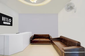 Uma área de recepção de hotel com balcão, um sofá de couro marrom e um ventilador fixado na parede.