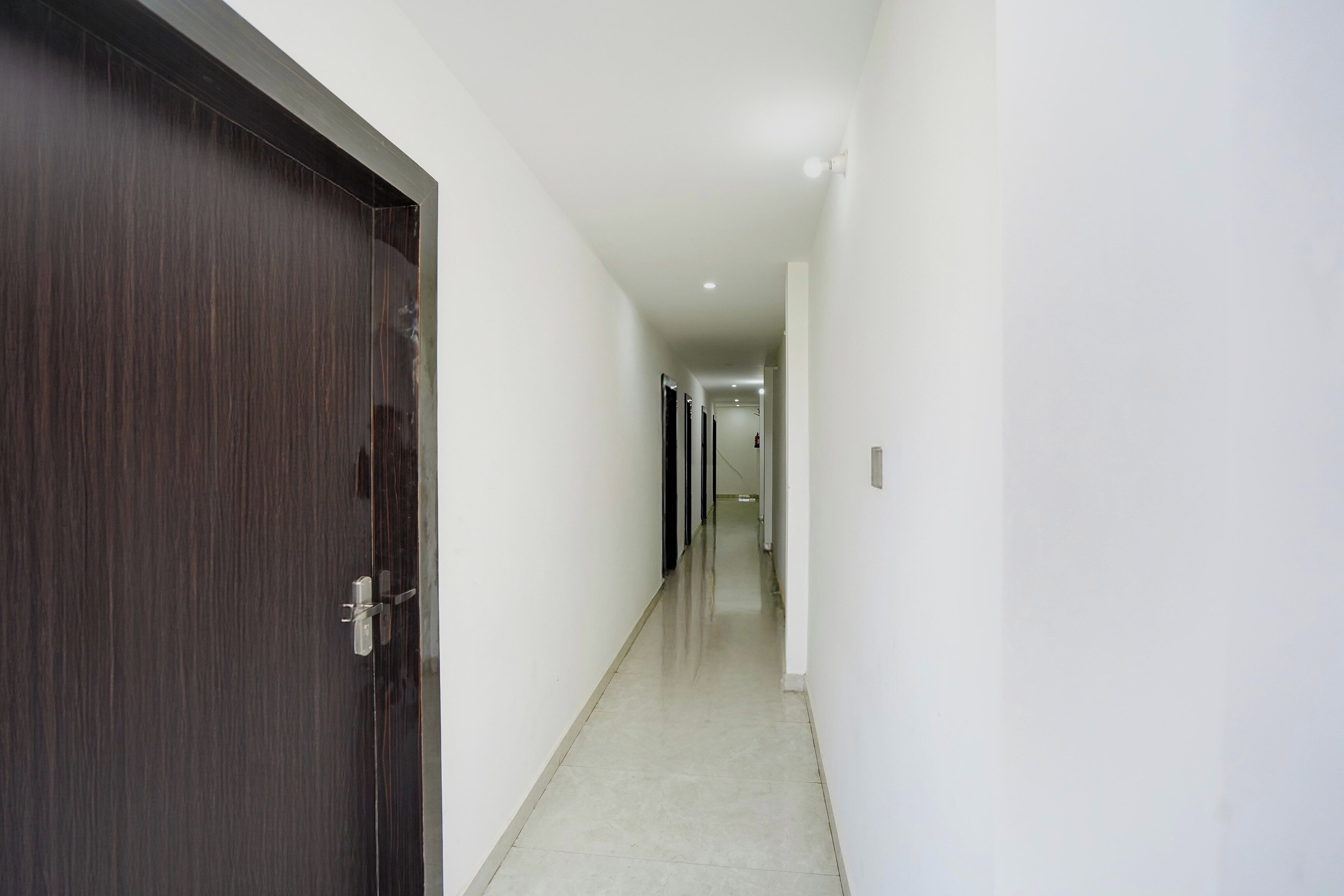 A corridor with a dark wooden door on the left, white walls, and a tiled floor.