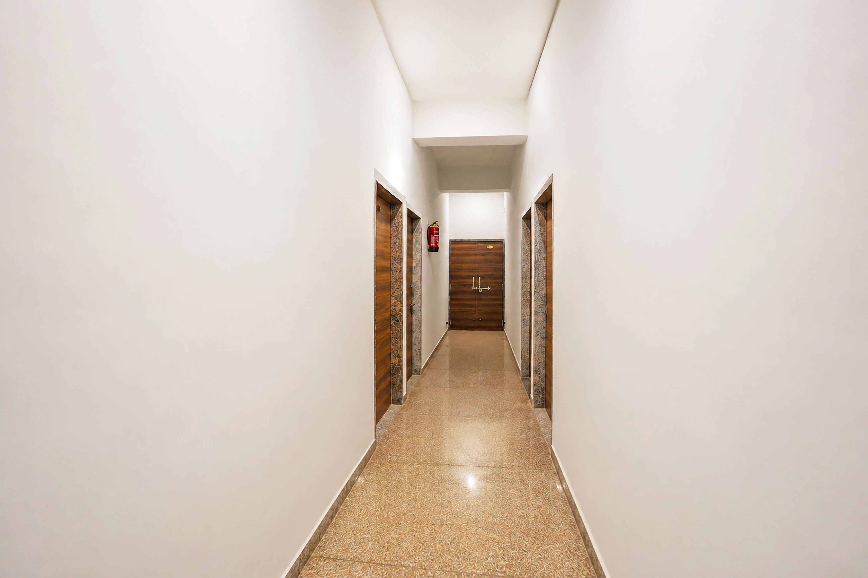 Un pasillo largo y estrecho con piso pulido, paredes blancas y puertas de madera a ambos lados.