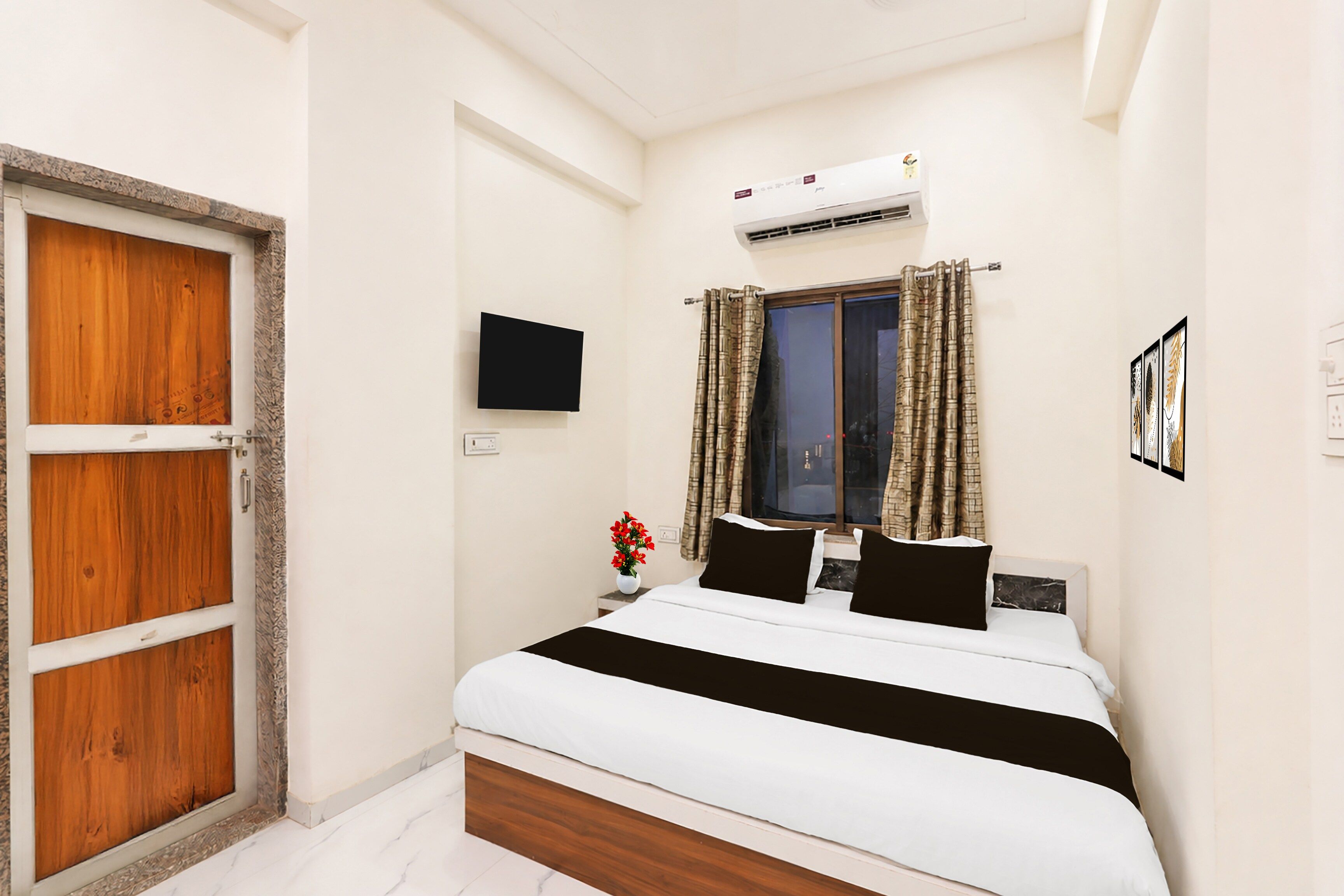 A bedroom with a bed, a wooden door, a wall-mounted TV, an air conditioner, and a window with curtains.
