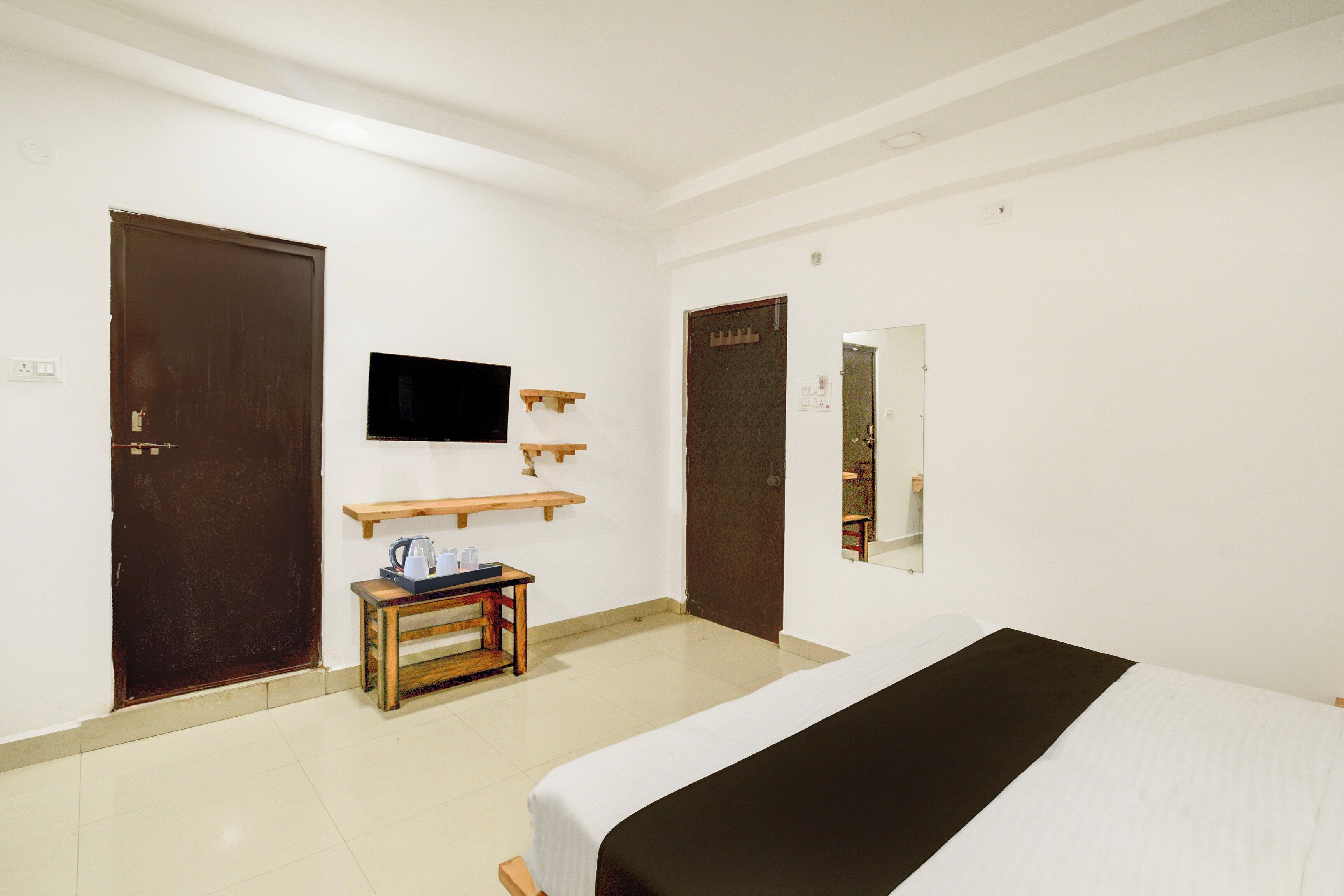 A hotel room with a bed, a television mounted on the wall, a wooden side table with a kettle, and two doors.