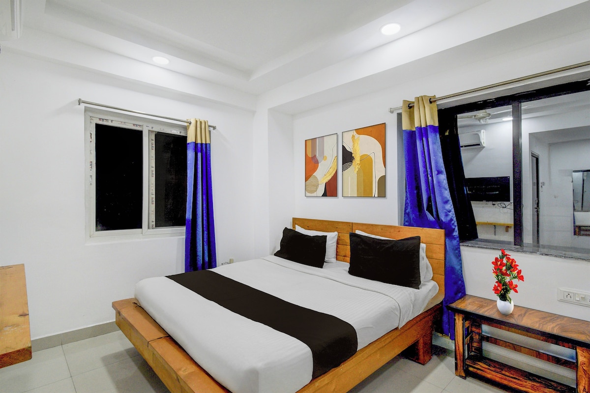 Une chambre à coucher avec un lit, une fenêtre, des rideaux, une table de chevet avec un vase, et des œuvres d’art accrochées au mur.