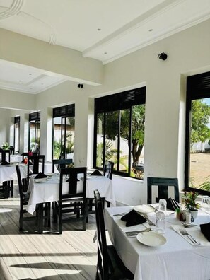 An indoor dining area with tables set for two, large windows, and a view of greenery outside.