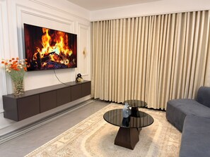 A living room with a flat-screen TV, a dark-colored sofa, a glass coffee table, a patterned rug, a vase with flowers, and a large window with beige curtains. - Bellace Haven | Stylish Holiday Homes & Short-Stay Accommodation in Kenya (Nairobi)