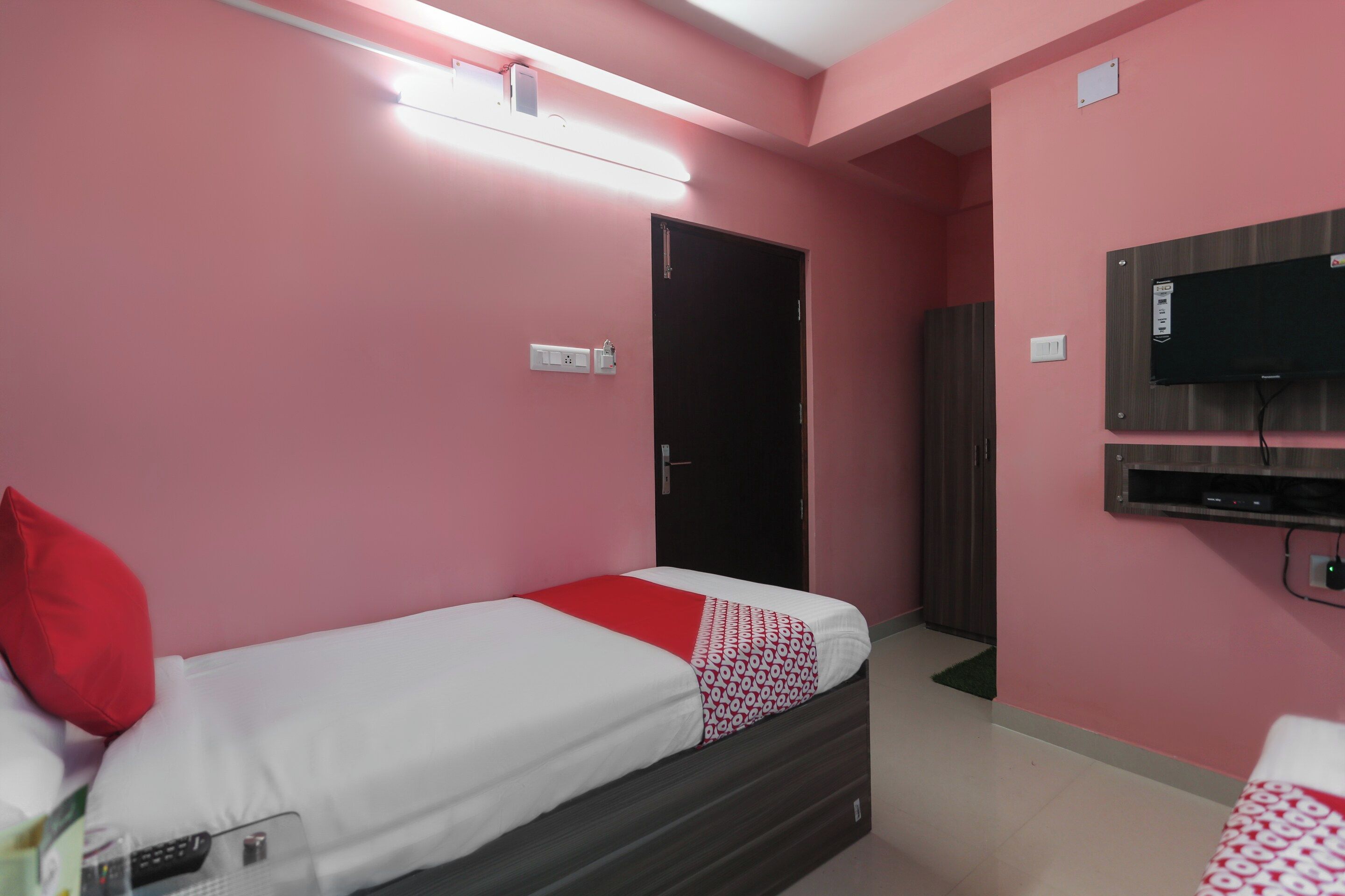 Una cama individual con una manta de diseño rojo y blanco, un televisor instalado en la pared, y una puerta que da a otra habitación.