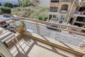 A balcony with a potted plant, a metal railing, and a view of a street with parked cars and buildings. - Spacious 2 Bed 2 Bath Apartment in Swieqi (Swieqi)