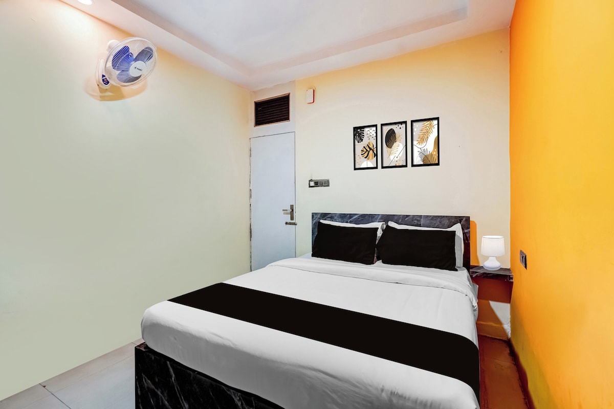 Un dormitorio con cama, mesita de noche, lámpara, ventilador de pared y dos cuadros enmarcados.
