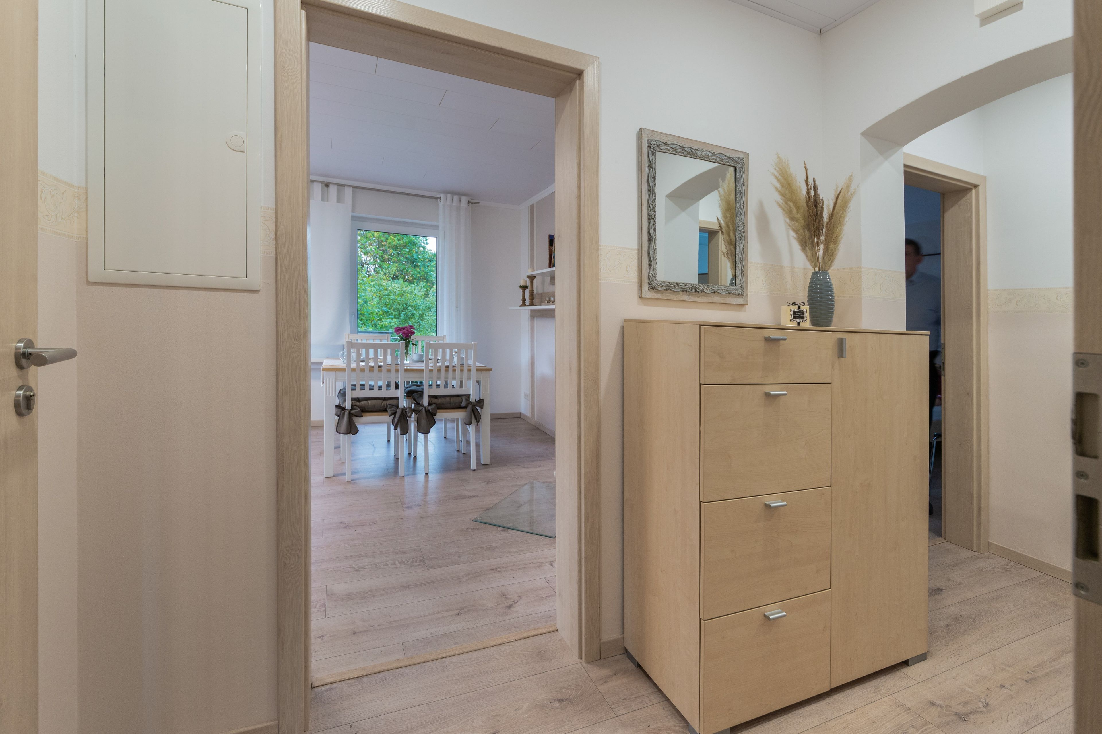 A hallway with a wooden wardrobe, a dining area with a table and chairs, and a window with a view of greenery.