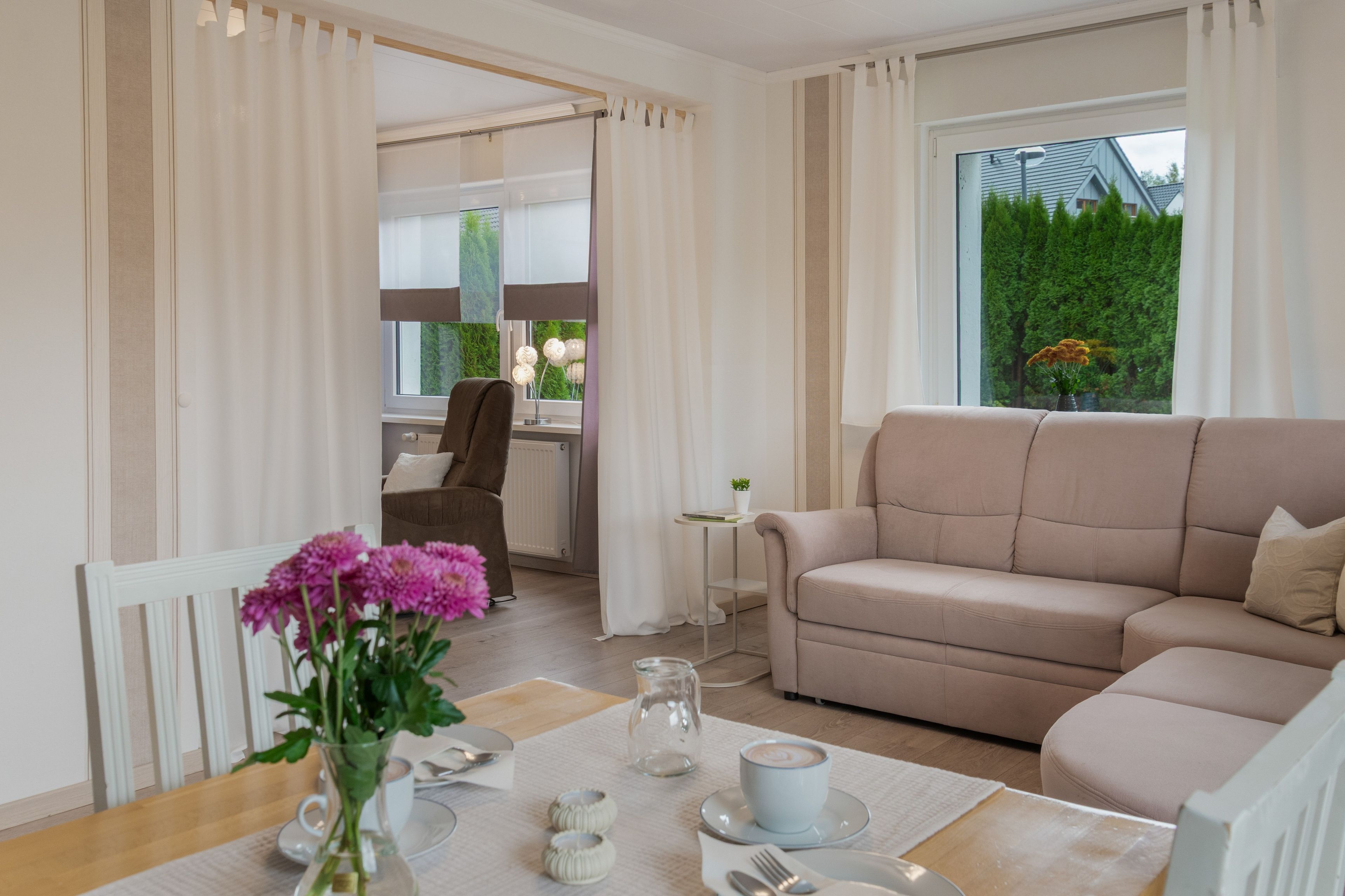 Ein modernes Wohnzimmer mit einem beigefarbenen Sofa, einem Esstisch mit Blumenvase und Blick auf eine grüne Landschaft durch das Fenster.