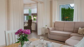 Ein modernes Wohnzimmer mit einem beigefarbenen Sofa, einem Esstisch mit Blumenvase und Blick auf eine grüne Landschaft durch das Fenster.