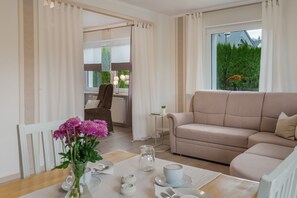 Ein modernes Wohnzimmer mit einem beigefarbenen Sofa, einem Esstisch mit Blumenvase und Blick auf eine grüne Landschaft durch das Fenster.