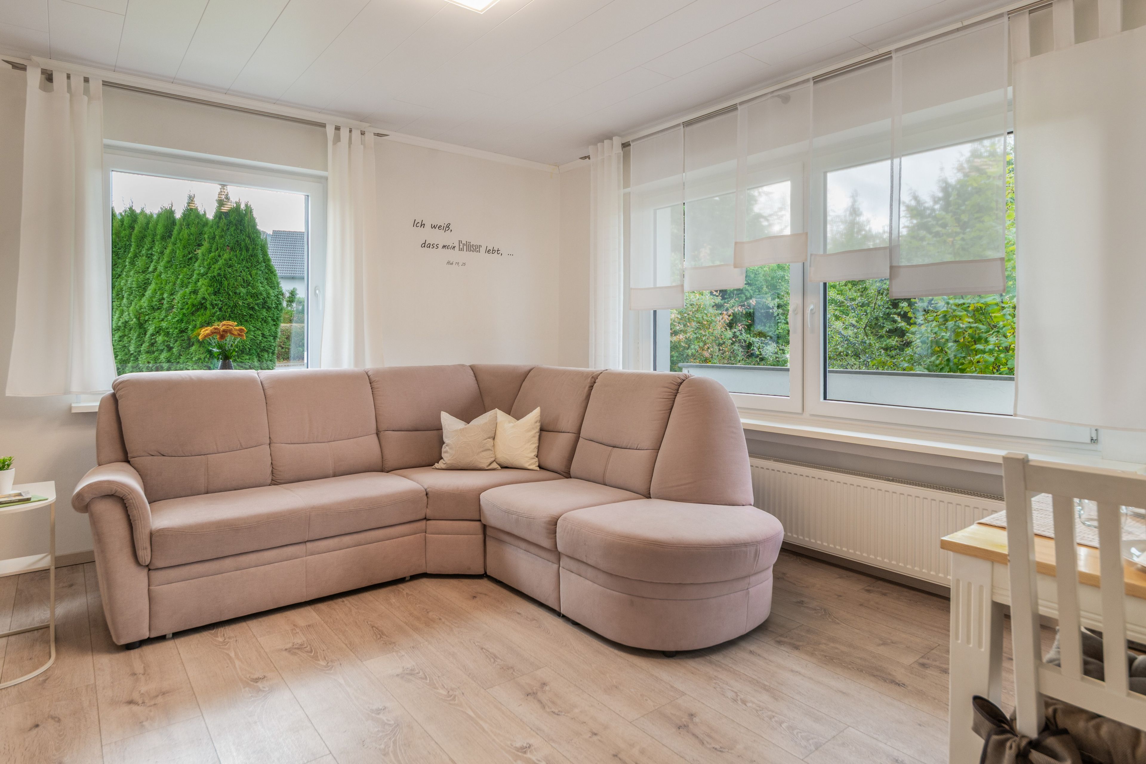 Ein modernes Wohnzimmer mit einem beigen Ecksofa, einem Esstisch und großen Fenstern mit Blick auf Grünflächen.