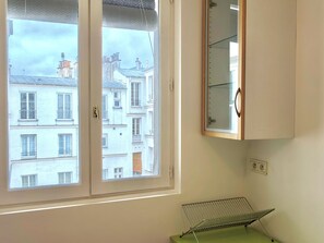 A room with a window overlooking a building, a metal drying rack, and a white cabinet with a mirror. - New Listing (Paris)