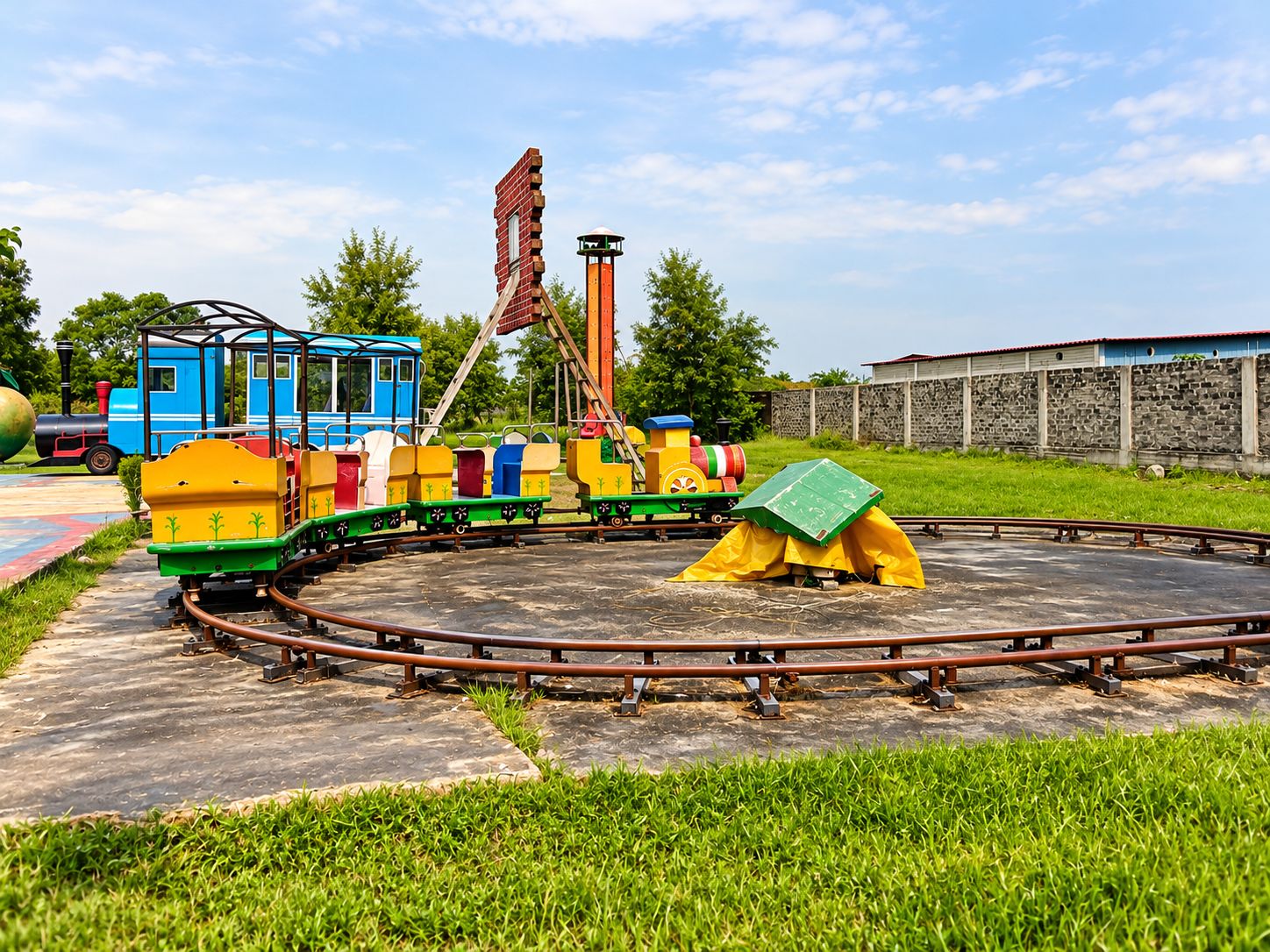 Un paseo en tren infantil colorido con una vía circular, un pequeño vagón azul y una estructura verde tipo tienda en el centro.